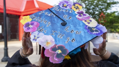A student with a graduation cap painted blue with tropical flowers with black letter reading "There's just no telling how far I'll go"